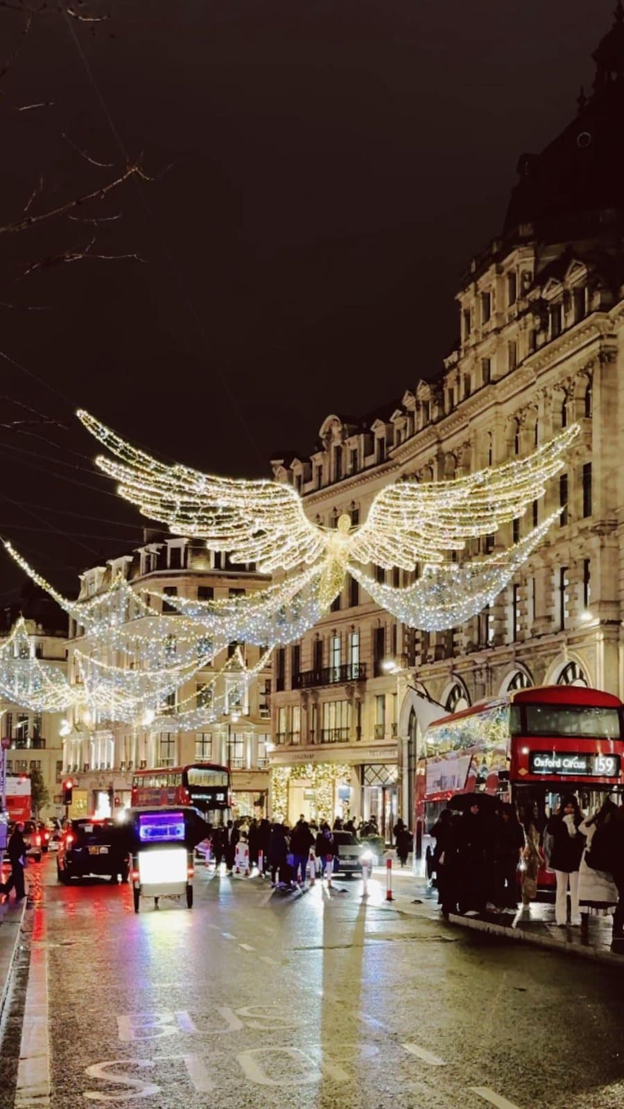 A London street at night under lights shaped like wings, with a red bus.